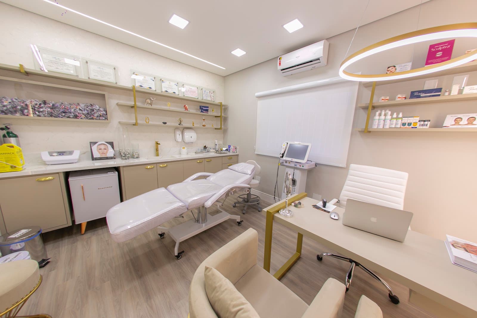 Sala de procedimentos com maca branca, bancada clara, prateleiras com perfis dourados e certificados na parede