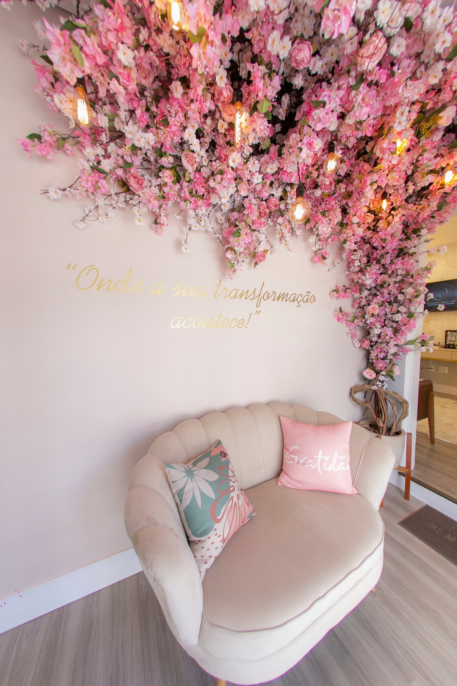 Poltrona clara sob um teto de flores cor-de-rosa e a frase dourada Onde a sua transformação acontece
